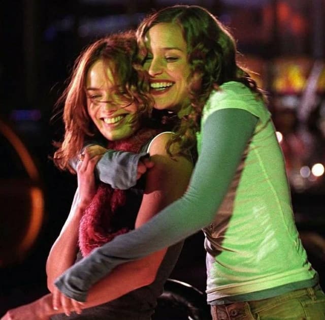 One woman seems to be hugging another, as they look into something. They both have warm clothes on. There's people behind them, from the movie one can know they're in a stadium.