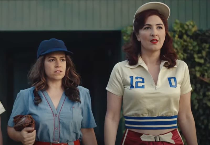 Two women dressed in baseball gear are looking at something behind the camera