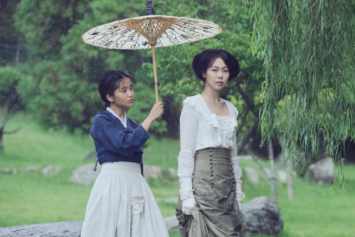 Two women are in a garden. One of them is dressed as a maid, and is holding an umbrella over the other one.